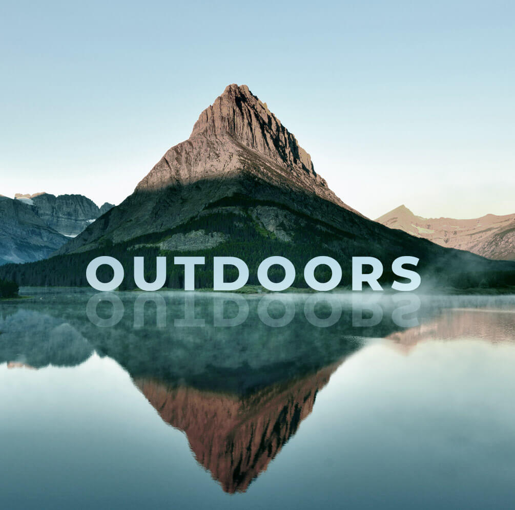 A large mountain within Glacier National Park is pictured with a reflective body of water sitting in front of it. The word “OUTDOORS” for mobile browsers